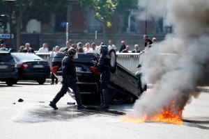 French riot police stand next to an overturned car as striking French taxi drivers demonstrate at the Porte Maillot to block the traffic on the Paris ring road during a national protest against car-sharing service Uber, in Paris, France, June 25, 2015. French taxi drivers stepped up protests against U.S. online cab service UberPOP on Thursday, blocking road access to airports and train stations in Paris and other cities. REUTERS/Charles Platiau  TPX IMAGES OF THE DAY